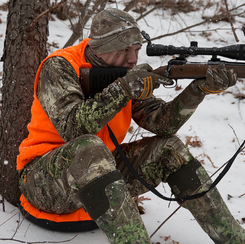 A hunter sitting under a tree aiming his firearm. Warm Up With the ThermaSeat Heat-A-Seat